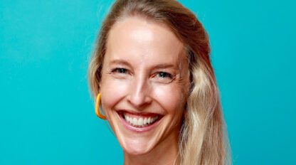 A woman with long blonde hair, wearing a black top and yellow earring, smiles at the camera in front of a blue background after completing the Boston Marathon.