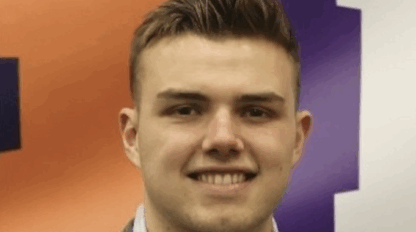A young man with short light brown hair wearing a grey blazer and light shirt stands in front of a purple and orange Boston Marathon background with large white letters.