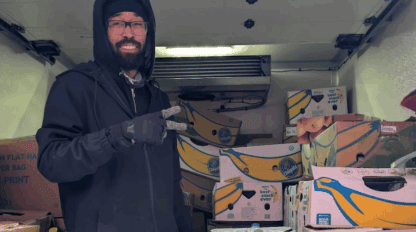 A man wearing glasses, a beanie, and gloves stands in a delivery truck filled with boxes of bananas, holding up a peace sign.