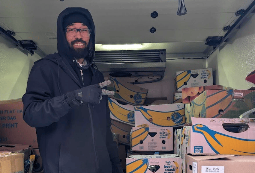 A man wearing glasses, a beanie, and gloves stands in a delivery truck filled with boxes of bananas, holding up a peace sign.