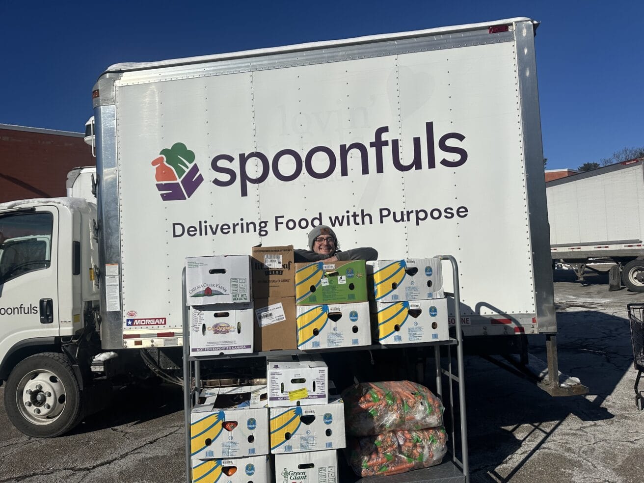 A person stands behind stacked food boxes in front of a "Lovin' Spoonfuls" truck with the slogan "Delivering Food with Purpose" in a parking lot.