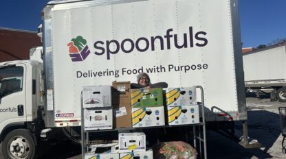 A person stands behind stacked food boxes in front of a "Lovin' Spoonfuls" truck with the slogan "Delivering Food with Purpose" in a parking lot.