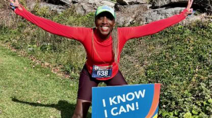 A runner in a red outfit kneels and poses with arms raised in front of a sign that reads "I KNOW I CAN!" along the Boston Marathon route, with trees and rocks dotting the grassy roadside in the background.