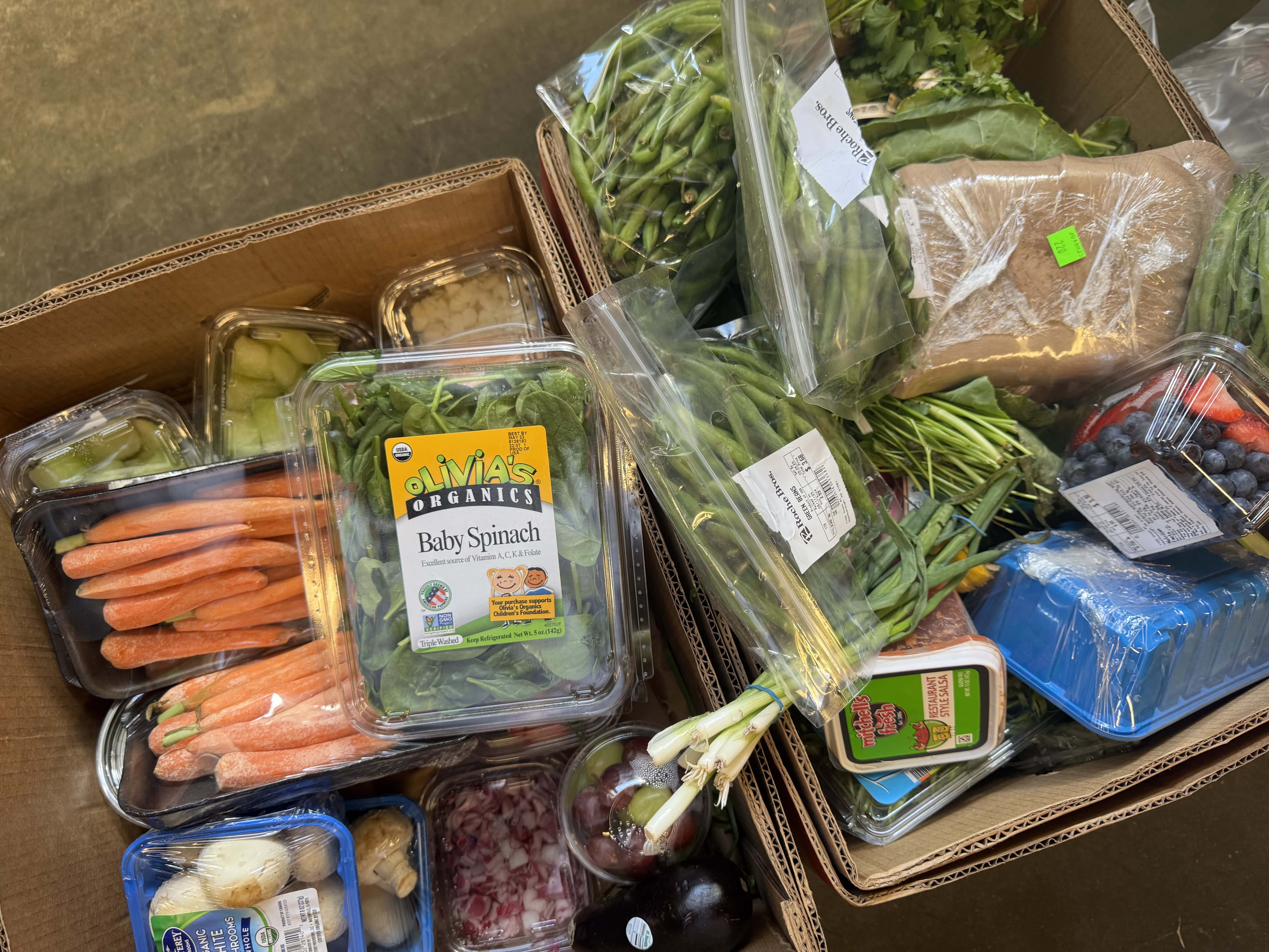 Two boxes filled with assorted groceries including fresh vegetables, greens, berries, packaged meat, and various plastic containers with produce.
