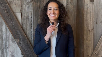 A person with curly hair wearing a navy blazer and white shirt is holding a small microphone, standing in front of a wooden wall.