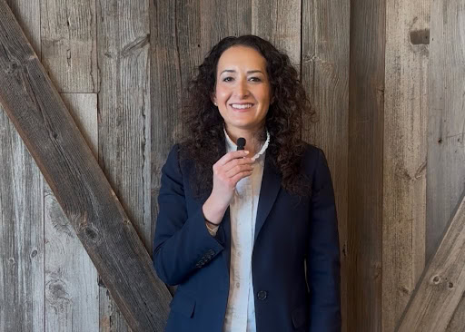 A person with curly hair wearing a navy blazer and white shirt is holding a small microphone, standing in front of a wooden wall.