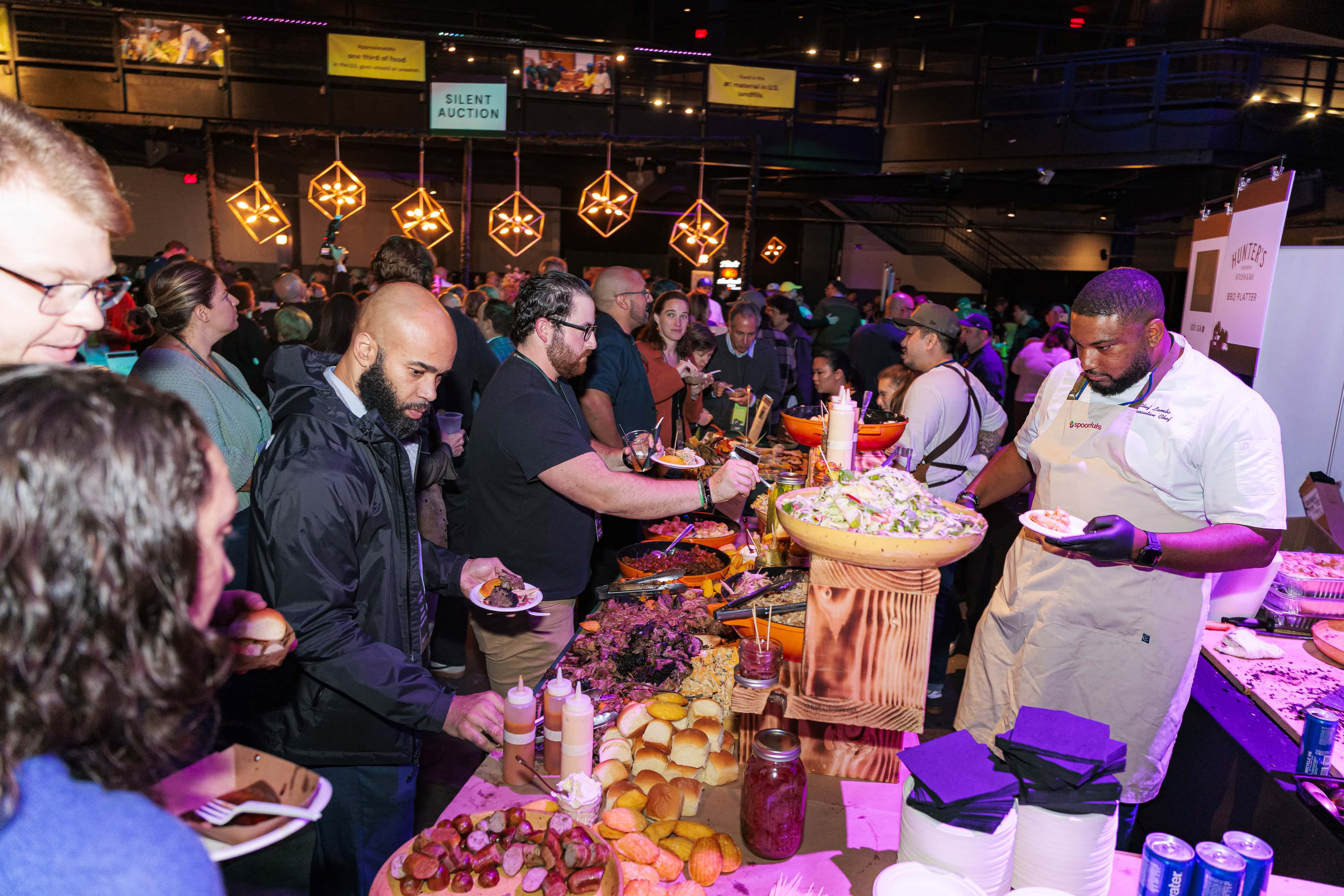 People serve themselves food from a buffet table at a crowded indoor event, with various dishes and salads displayed and a chef assisting on the right.