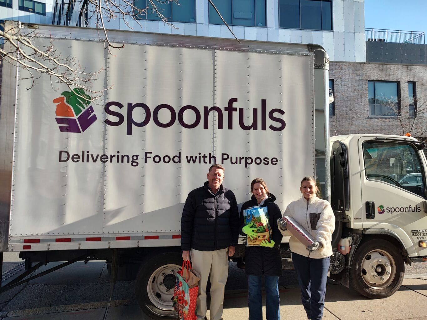 Three people stand in front of a Spoonfuls truck labeled "Delivering Food with Purpose," each holding bags or boxes, reflecting the organization’s Core Values & Inclusion Tenets on a sunny day in an urban area.
