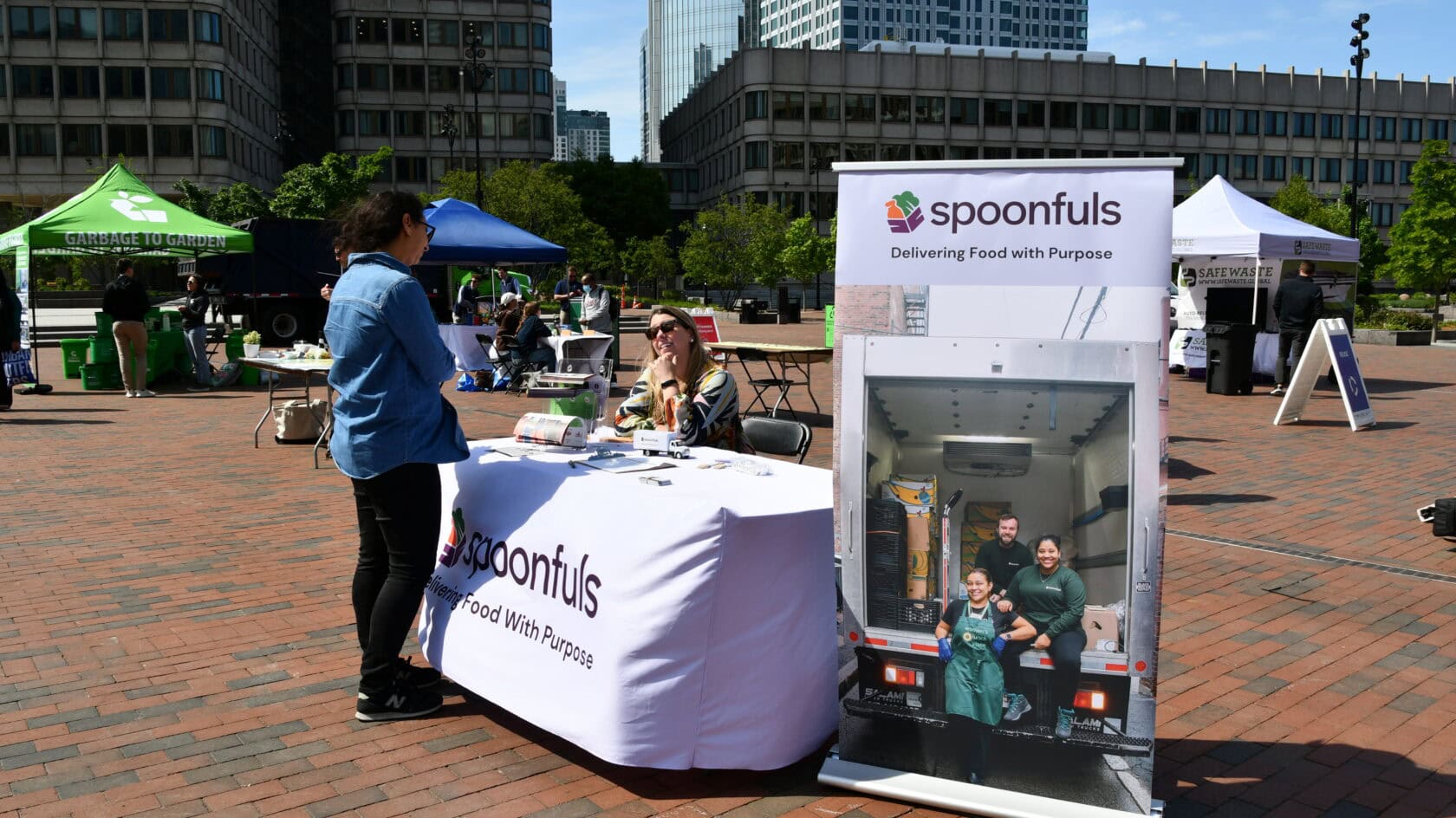 A person speaks with a representative at a Spoonfuls booth in an outdoor plaza, with a large banner displaying "Delivering Food with Purpose" and a photo of a delivery truck.