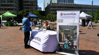 A person speaks with a representative at a Spoonfuls booth in an outdoor plaza, with a large banner displaying "Delivering Food with Purpose" and a photo of a delivery truck.