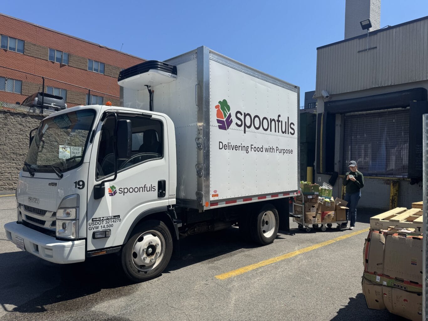A Spoonfuls delivery truck is parked next to a loading dock as a worker, guided by our Core Values & Inclusion Tenets, unloads boxes of food onto carts.