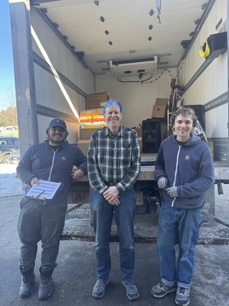 Three men stand at the back of an open delivery truck—two in hoodies and gloves, one in a plaid shirt—working together with boxes and supplies, demonstrating our Core Values & Inclusion Tenets in action.