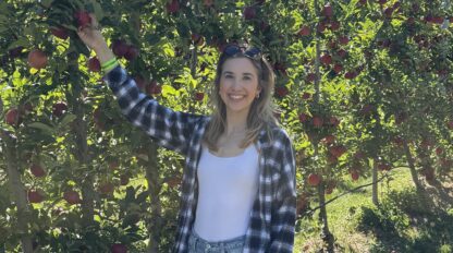 A woman in a plaid shirt and jeans stands outdoors, reaching for an apple on a tree in an orchard on a sunny day, wearing her Boston Marathon cap with pride.