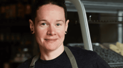 A person wearing a dark shirt and apron smiles while standing indoors, with shelves and a display case in the background, proudly sporting a Boston Marathon pin.
