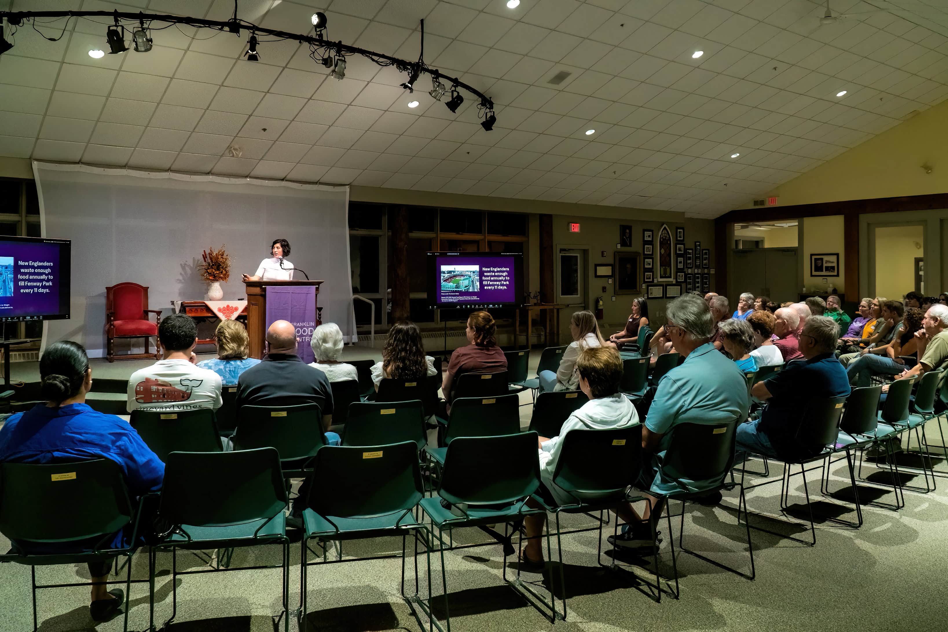 A speaker presents at a podium to an audience seated in a room, with two screens displaying slides on either side of the stage.