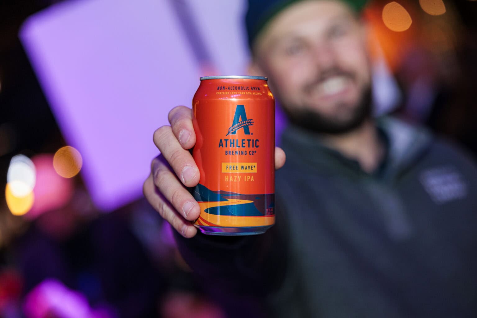 A person holds an orange can of Athletic Brewing Co. Free Wave Hazy IPA, perfect for your Ultimate Tailgate, with the background softly blurred.