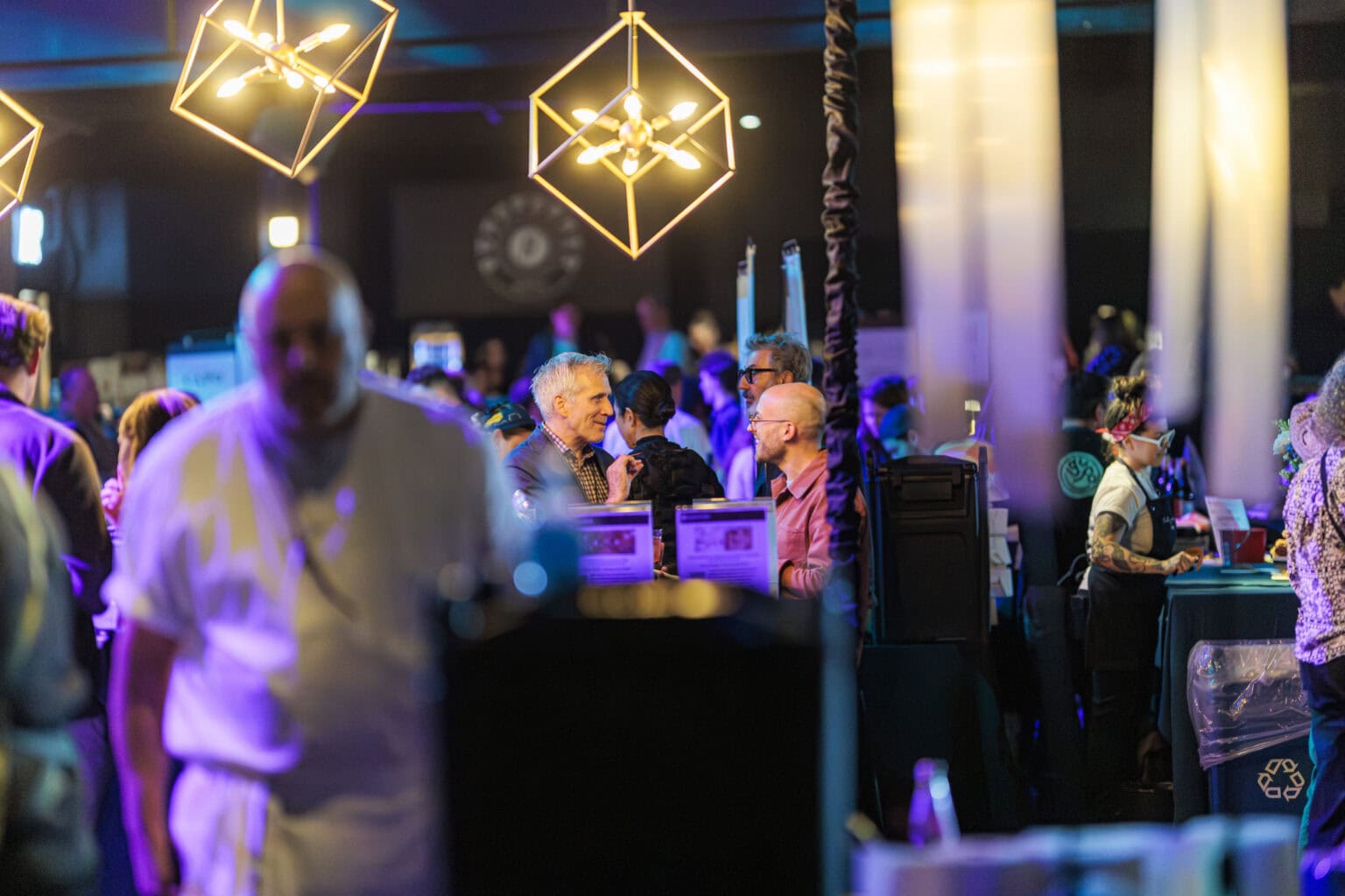 A group of people socialize under geometric pendant lights at an indoor Ultimate Tailgate event, illuminated by vibrant blue and purple lighting.