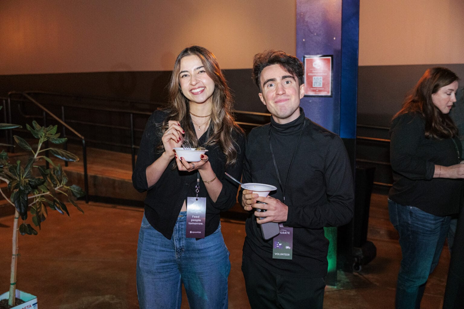 Two people stand indoors at the Ultimate Tailgate event, smiling and holding bowls of food, both wearing lanyards. Another person stands in the background near a plant.