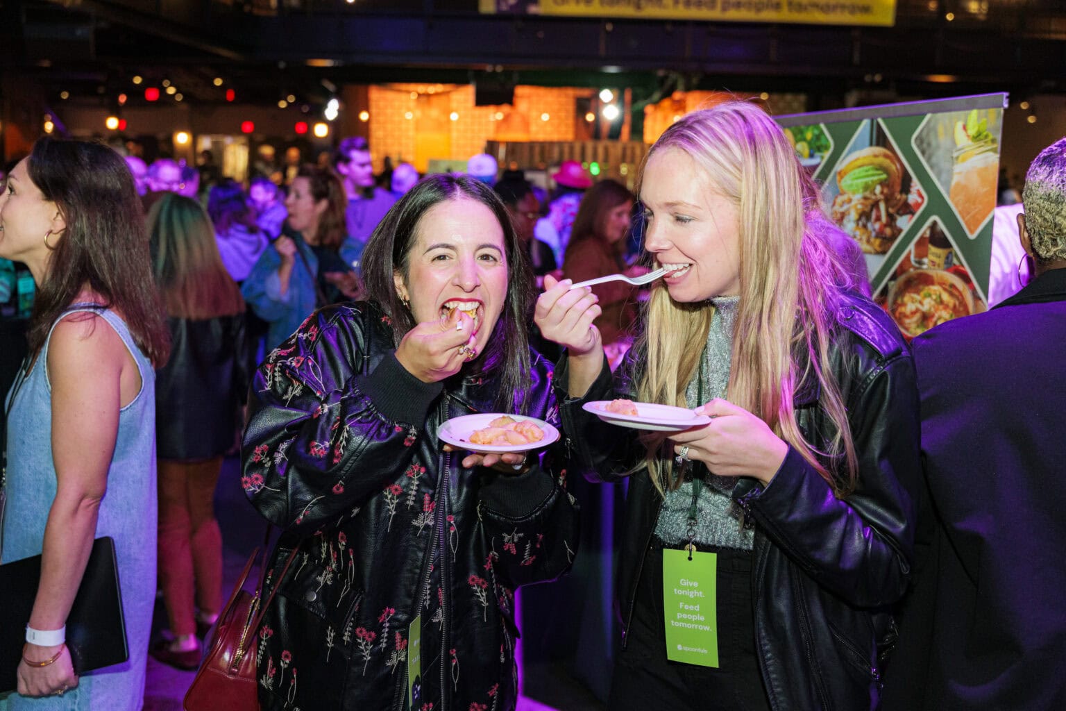 Two women stand in a crowded event space at the Ultimate Tailgate, smiling and enjoying delicious bites from their plates. Other attendees and impressive food displays are visible in the background under colorful lighting.