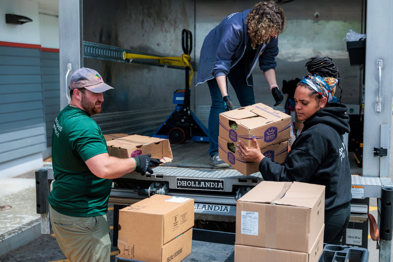 Three people unload cardboard boxes from a truck, stacking them on the platform at the back of the vehicle.