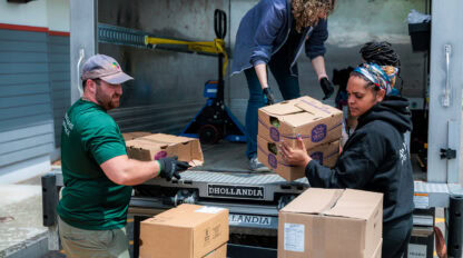 Three people unload cardboard boxes from a truck, stacking them on the platform at the back of the vehicle.