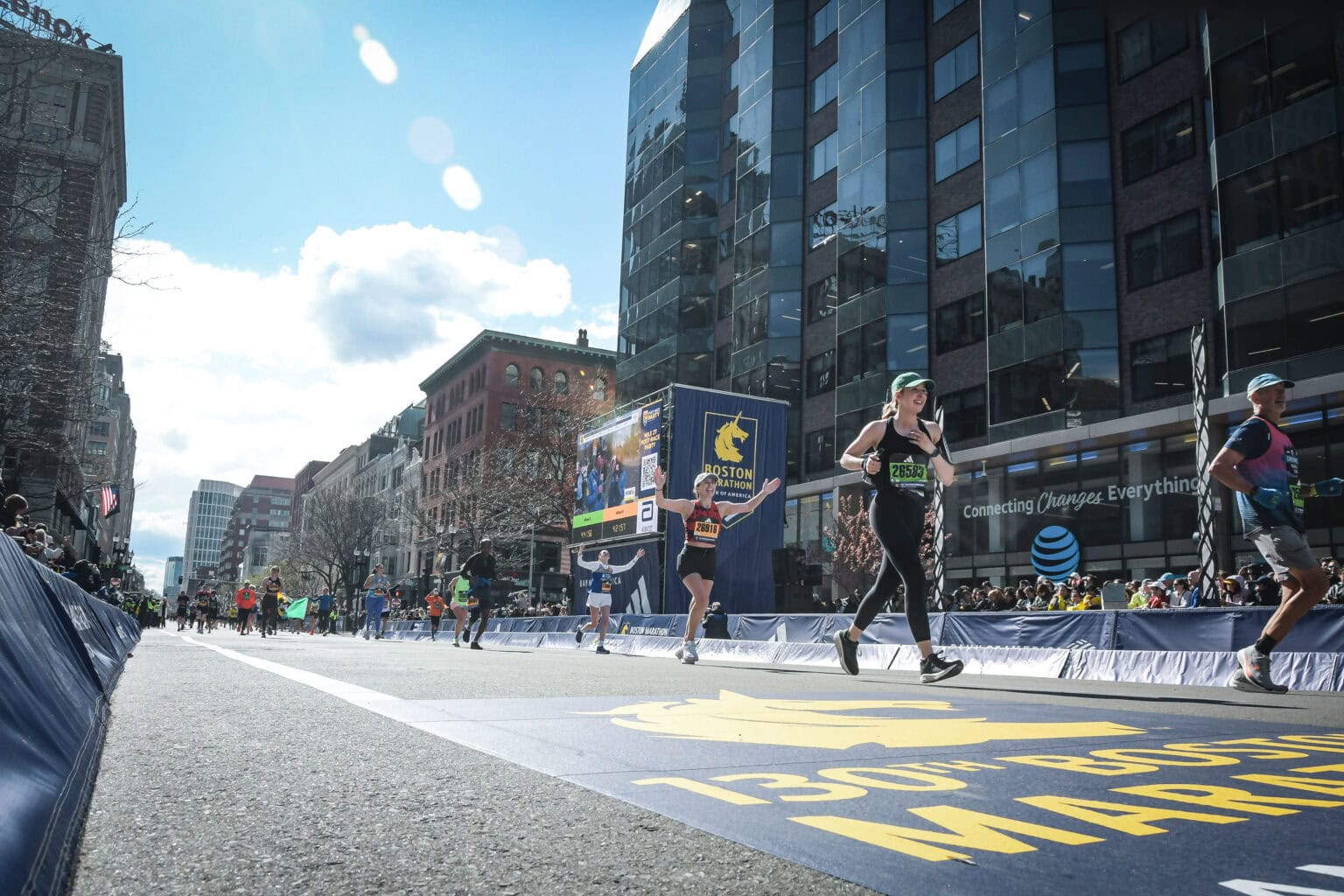 Runners in athletic gear compete in the Boston Marathon on a sunny day, with spectators and tall glass buildings in the background, creating an atmosphere as lively and energetic as top Events like the Battle of the Bites.