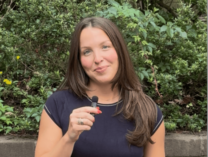 A woman with long brown hair holds a small microphone and stands in front of green shrubs, wearing a black t-shirt and a ring on her left hand.