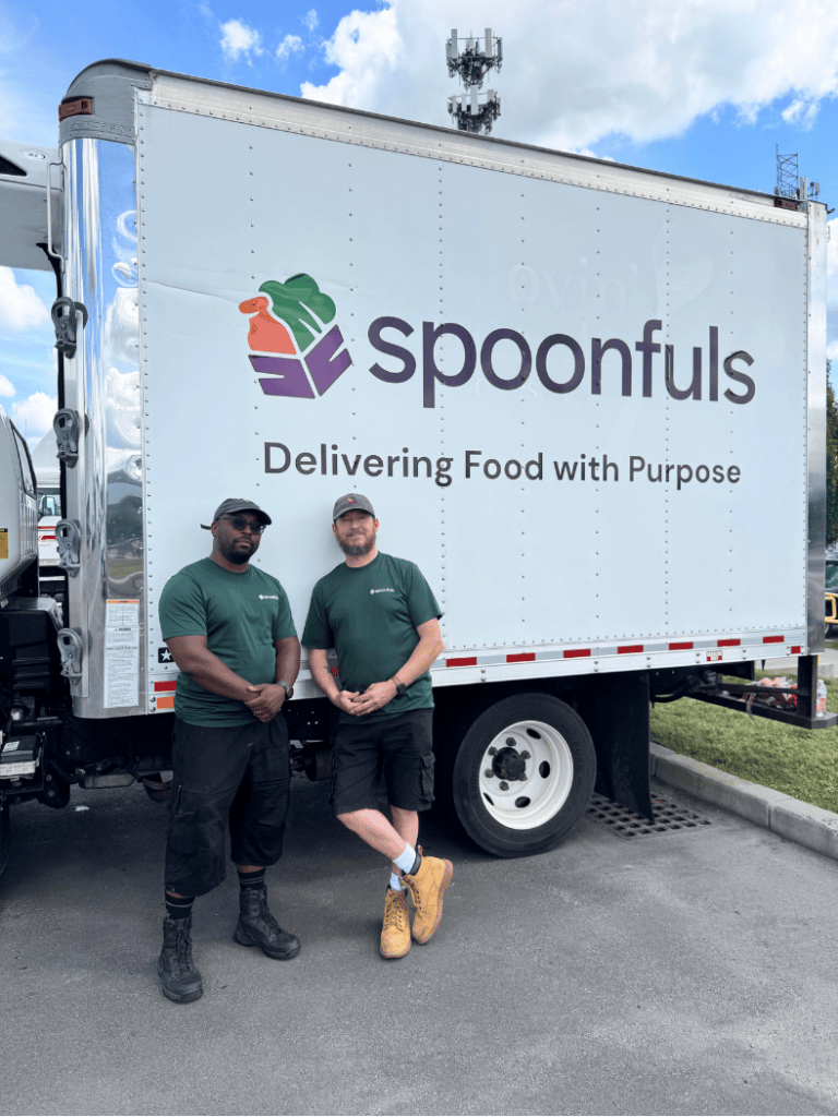 Two employees in branded green t-shirts stand in front of a white box truck with the “Spoonfuls” logo and the text “Delivering Food with Purpose.”