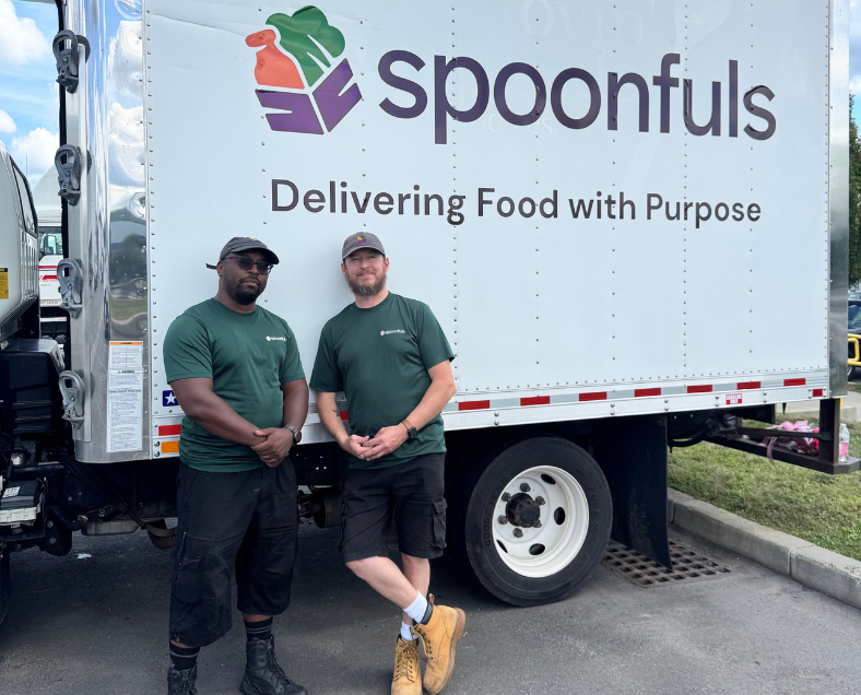 Two employees in branded green t-shirts stand in front of a white box truck with the “Spoonfuls” logo and the text “Delivering Food with Purpose.”