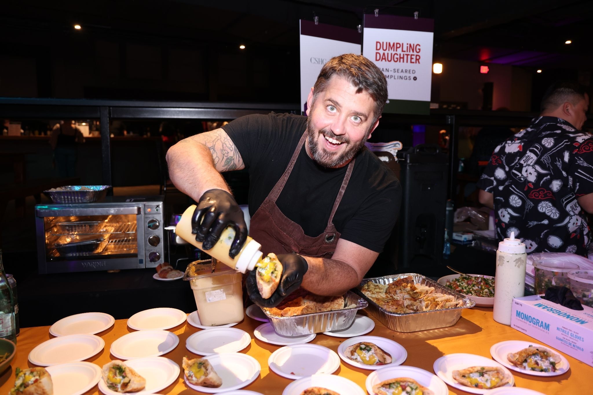 A smiling man wearing black gloves drizzles sauce onto a dumpling held over a metal food tray at a Dumpling Daughter station.