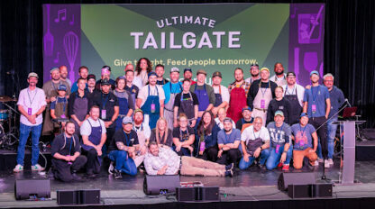 A large group of people posing for a photo on a stage in front of an “Ultimate Tailgate” event banner.