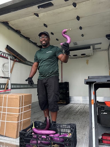 A man standing inside the back of a delivery truck holds up a curved purple eggplant.